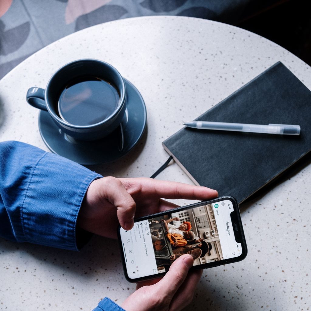 The Pressure of Pretending You're Okay A person views Instagram on a smartphone at a cafe table with coffee, notebook, and pen.
